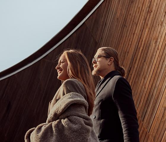 Man with long hair and glasses and a women with a grey coat are looking away to the left